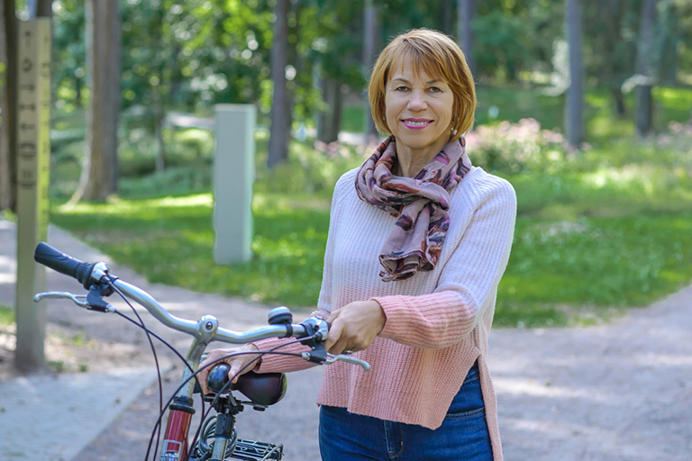 Natural Pharmaceuticals „VitaSolaris® D3“ Vidutinio amžiaus moteris gamtoje stovi su raudonu dviračiu ir dėvi rožinį megztinį bei šaliką.