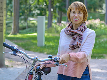 Natural Pharmaceuticals „VitaSolaris® D3“ Moteris su džemperiu ir šaliu laikosi dviratį parke saulėtoje aplinkoje.