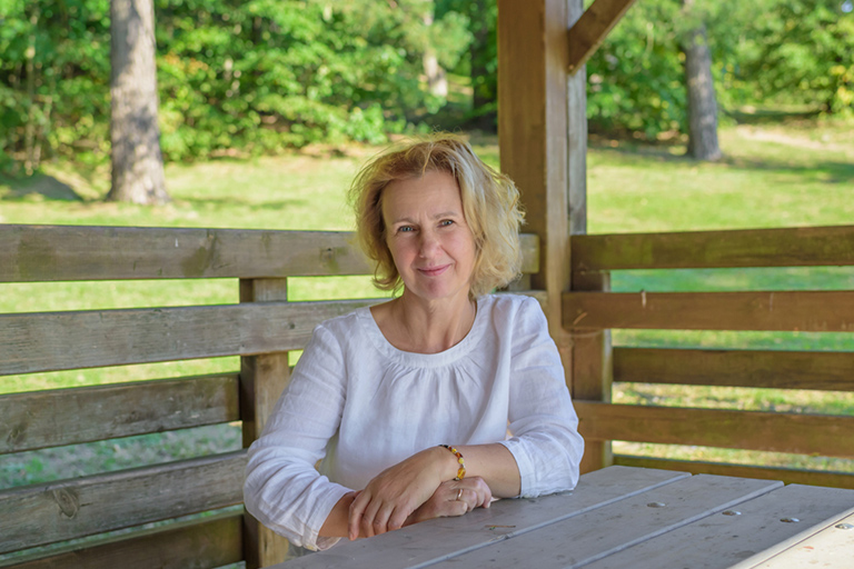 Natural Pharmaceuticals „VitaSolaris® D3“ Vidutinio amžiaus moteris su šviesiais plaukais sėdi poilsio pavėsinėje gamtoje, dėvi baltą palaidinę.