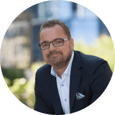 Natural Pharmaceuticals „VitaSolaris® D3“ Verslininkas dėvi kostiumą ir akinius, fone matomas miesto peizažas su pastatais ir žaluma.